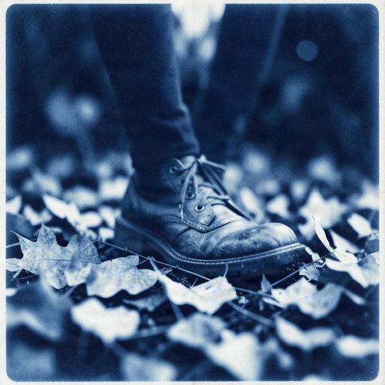 Cover art for Then And Now: a worn leather boot stepping through fallen leaves, in blue monochrome